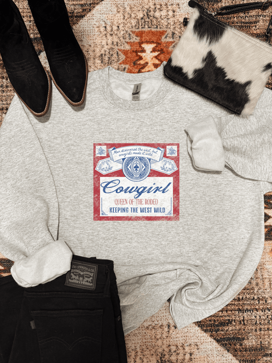 Flatlay of the Queen of the Rodeo Crewneck by Hides & Kinds, featuring a vintage red, white, and blue cowgirl graphic that reads "Man discovered the West, but cowgirls made it wild – Queen of the Rodeo – Keeping the West Wild," styled with black jeans, pointed boots, and a cowhide clutch on a western rug.