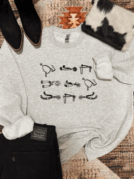 Ash grey crewneck with black western spurs design laid out with black jeans, cowhide purse, and black boots
