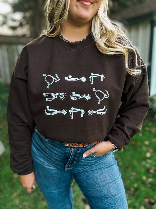 Woman wearing chocolate brown crewneck with white western spurs graphic, styled with jeans and belt