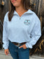 Woman wearing athletic grey quarter zip with turquoise Stampede logo by Hides & Kinds, standing in front of wood barn wall