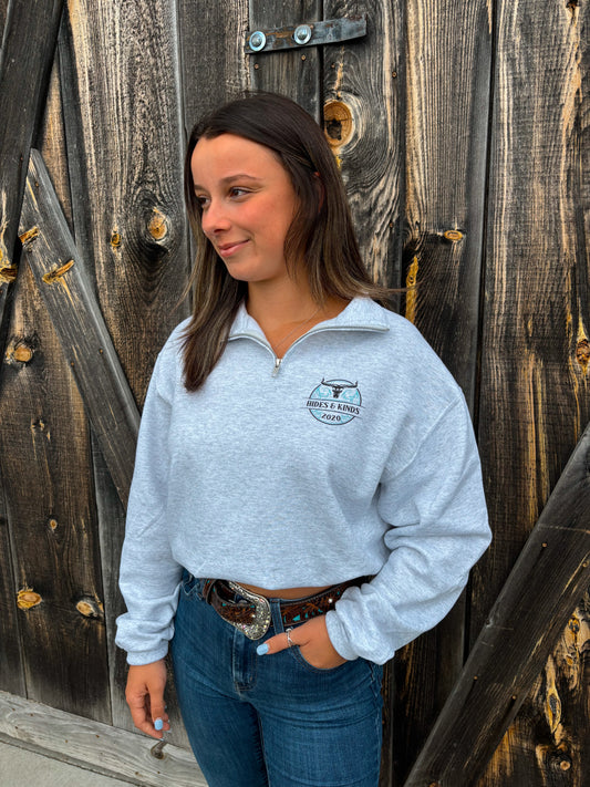 Full-body view of woman in light grey quarter zip with turquoise and black longhorn logo, styled with denim and western belt