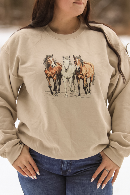 Model wearing the Trail Trio Crewneck standing in the snow with three galloping horses printed on the front, featuring a neutral sand colored sweatshirt perfect for cozy western style lovers.