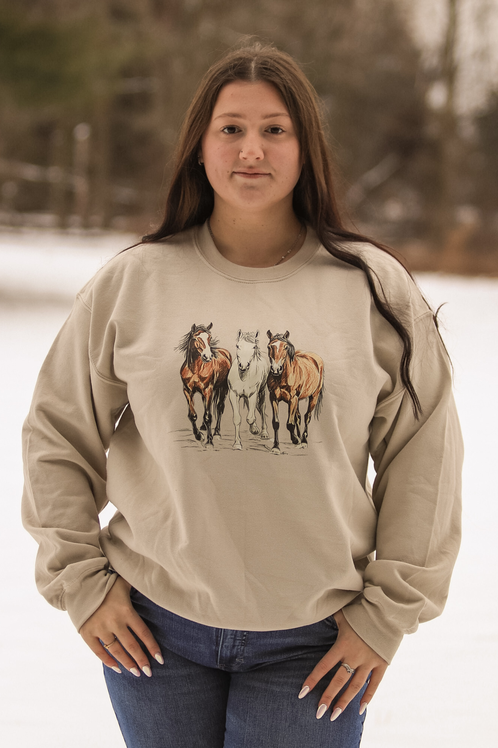Front facing photo of model styled in the Trail Trio Crewneck from Hides & Kinds, showcasing detailed artwork of three horses running together.