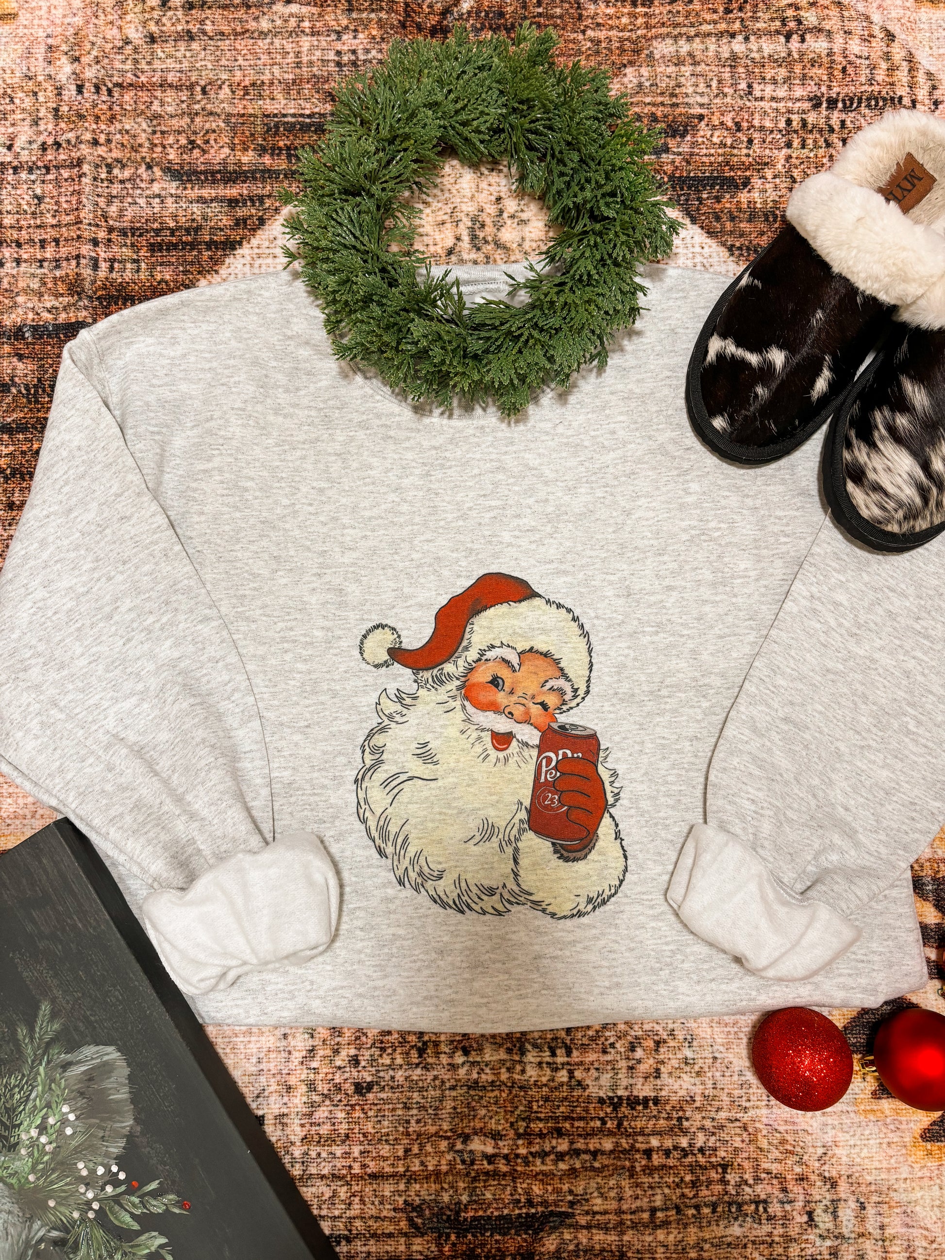 Flat lay of the Vintage Santa Dr Pepper Crewneck folded with holiday decor, showing retro Santa graphic holding a Dr Pepper can.
