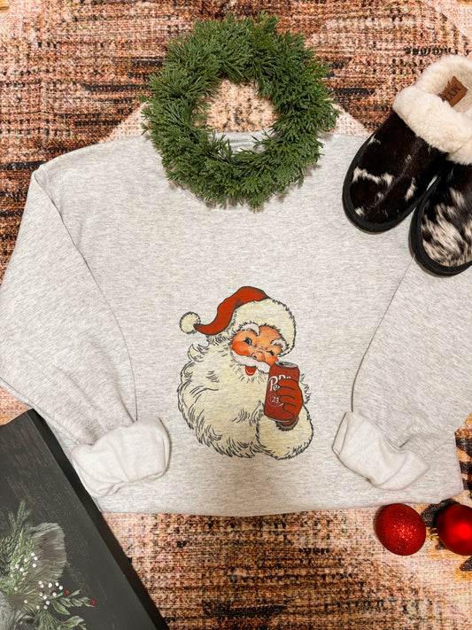 Flat lay of the Vintage Santa Dr Pepper Crewneck folded with holiday decor, showing retro Santa graphic holding a Dr Pepper can.
