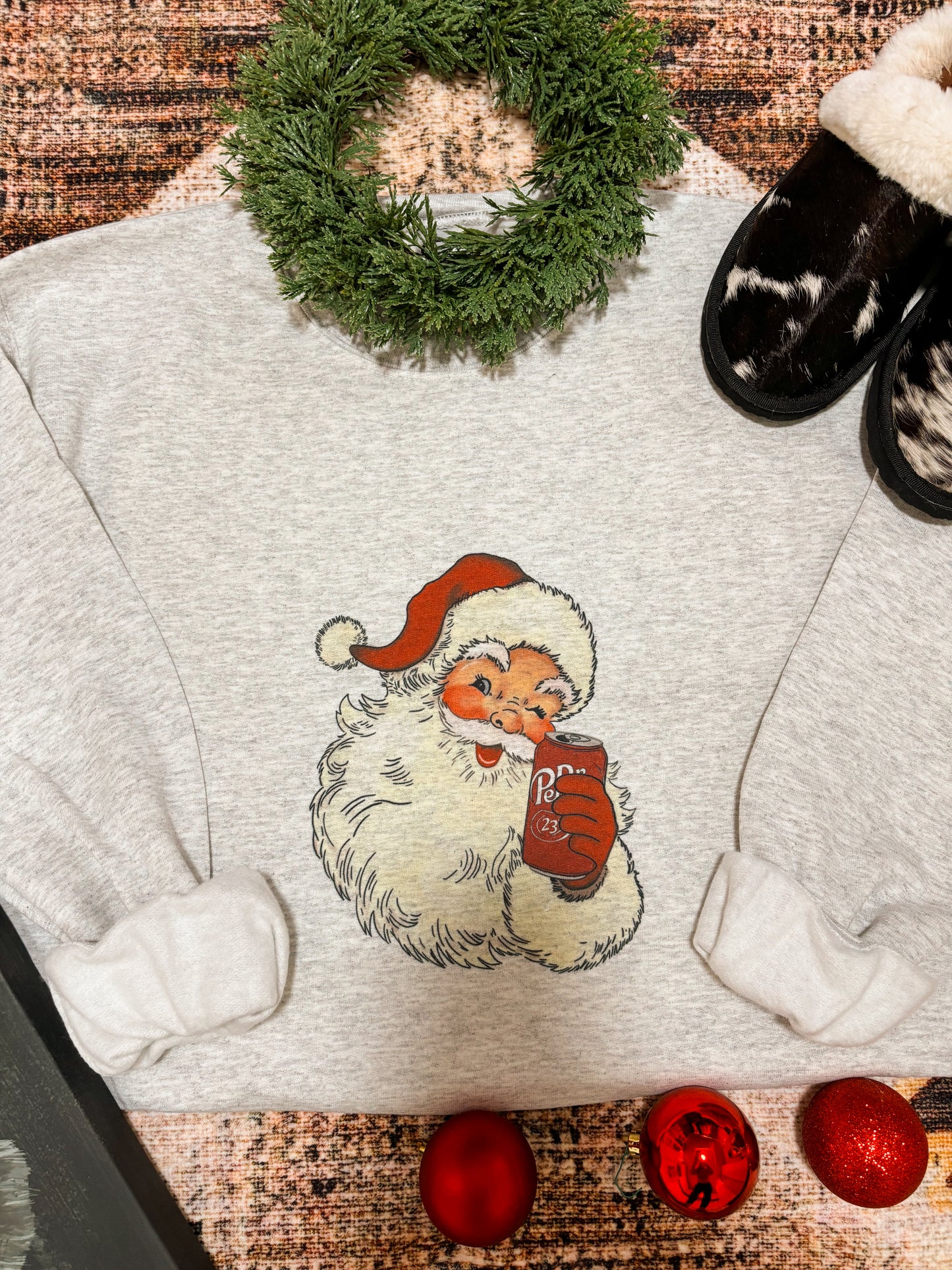 Close up of festive crewneck with retro Santa and Dr Pepper can graphic, styled with red ornaments and cozy textures.