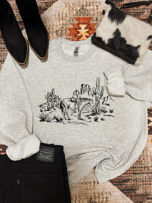 Flat lay of the Wild Horse Crewneck by Hides & Kinds, light grey sweatshirt with desert horse and cactus scene, styled with cowhide clutch and black denim