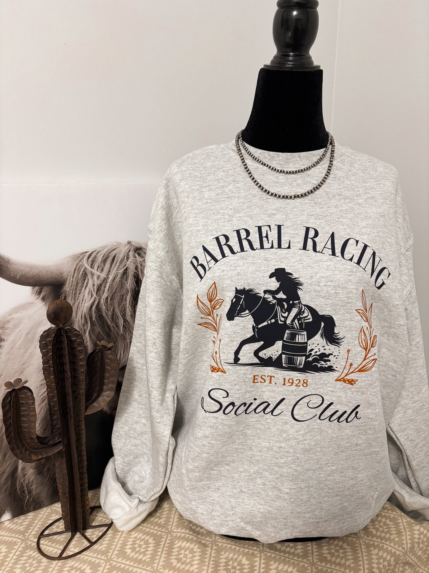 Western inspired grey crewneck with bold barrel racing design, styled beside a decorative cactus and rustic décor.