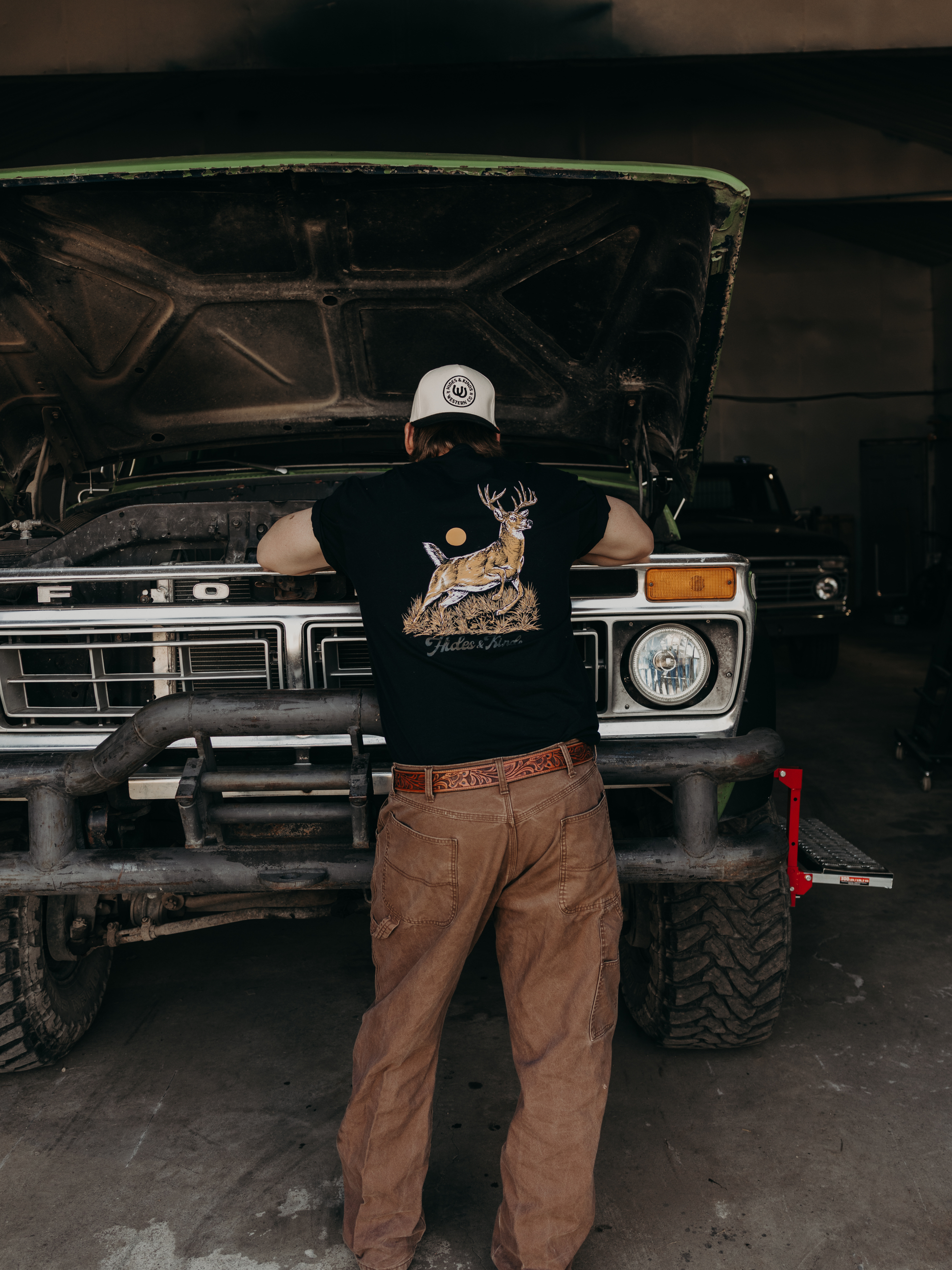 Model wearing the Hides & Kinds Big Buck Tee featuring a deer graphic while standing in front of a vintage truck.