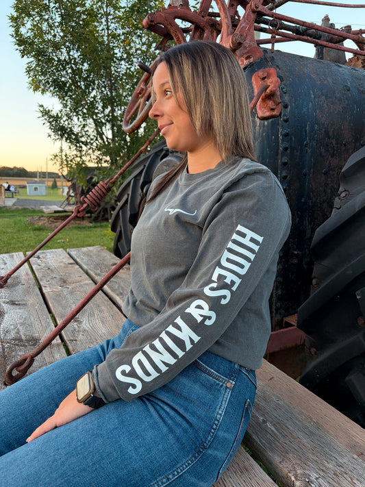 Model wearing the pepper grey Hides & Kinds Branded Longsleeve with white lettering down the sleeve and a white longhorn chest logo, seated beside vintage farm machinery.