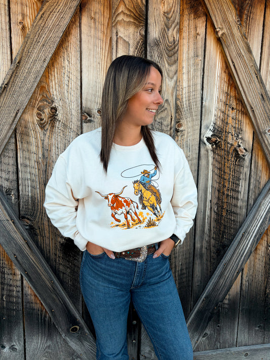 Model wearing the cream Hides & Kinds Breakaway Crewneck with a western cowboy roping graphic, styled with blue jeans in front of a rustic wooden barn.