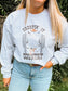 Woman wearing a light grey crewneck that says "Creepin' It Country" with a ghost in cowgirl boots and hat standing between two cacti.