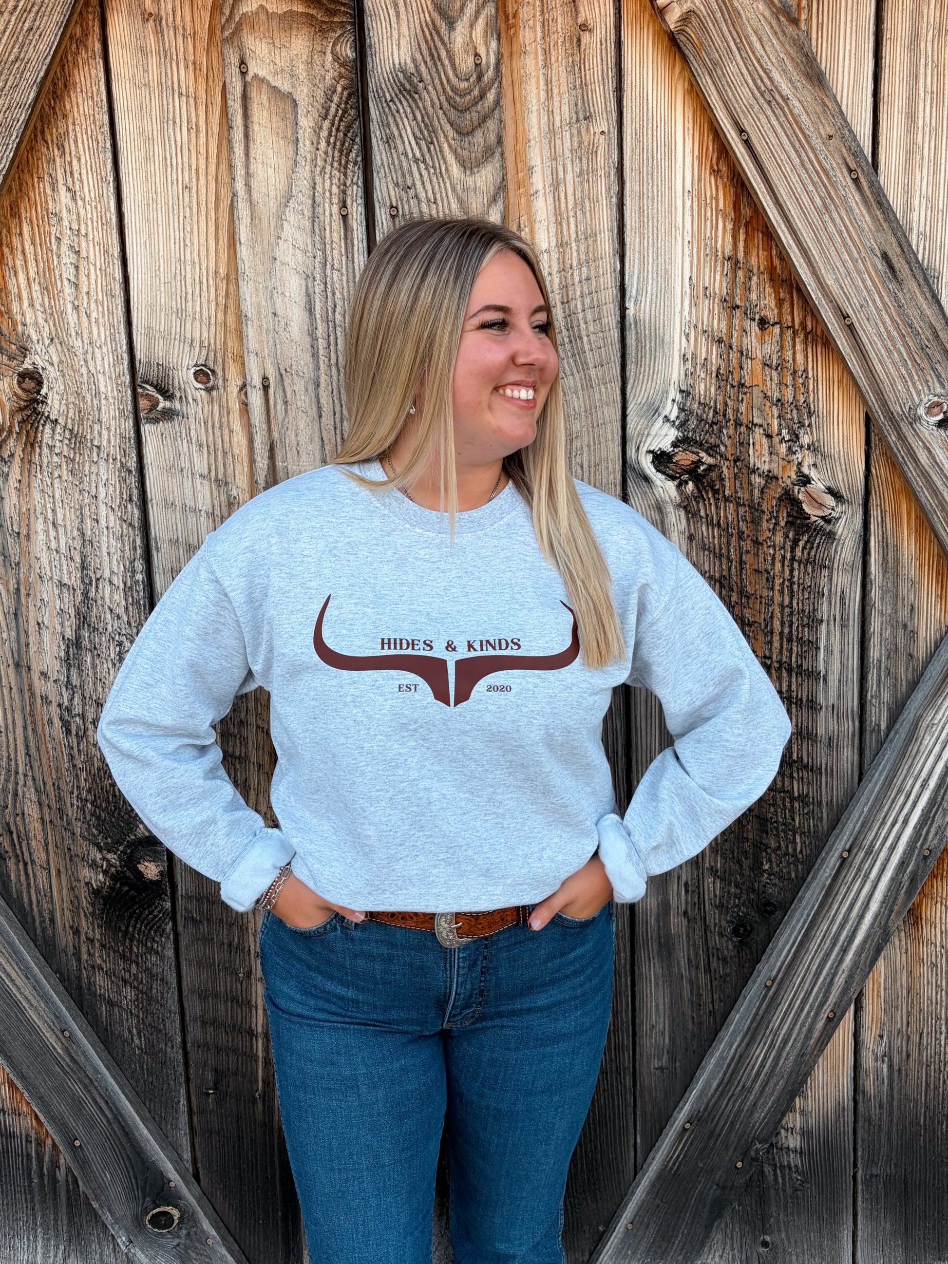 Model wearing the Hides & Kinds Original Logo Crewneck in ash grey with a brown longhorn graphic, paired with blue jeans in front of a rustic wooden barn door.