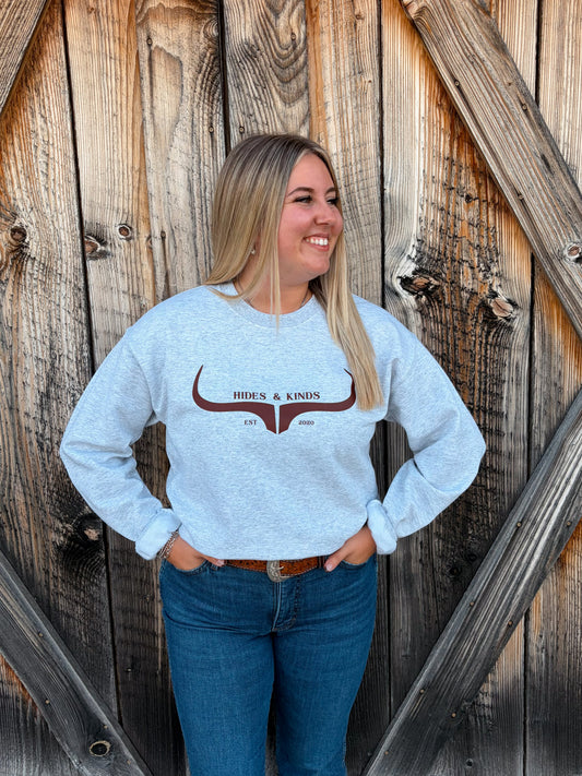 Model wearing the Hides & Kinds Original Logo Crewneck in ash grey with a brown longhorn graphic, paired with blue jeans in front of a rustic wooden barn door.