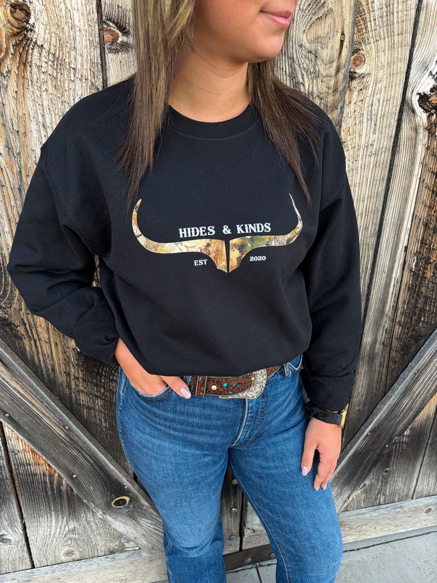 Close-up of Hides & Kinds Original Logo Crewneck in black with a camouflage longhorn graphic, styled with blue jeans and a western belt