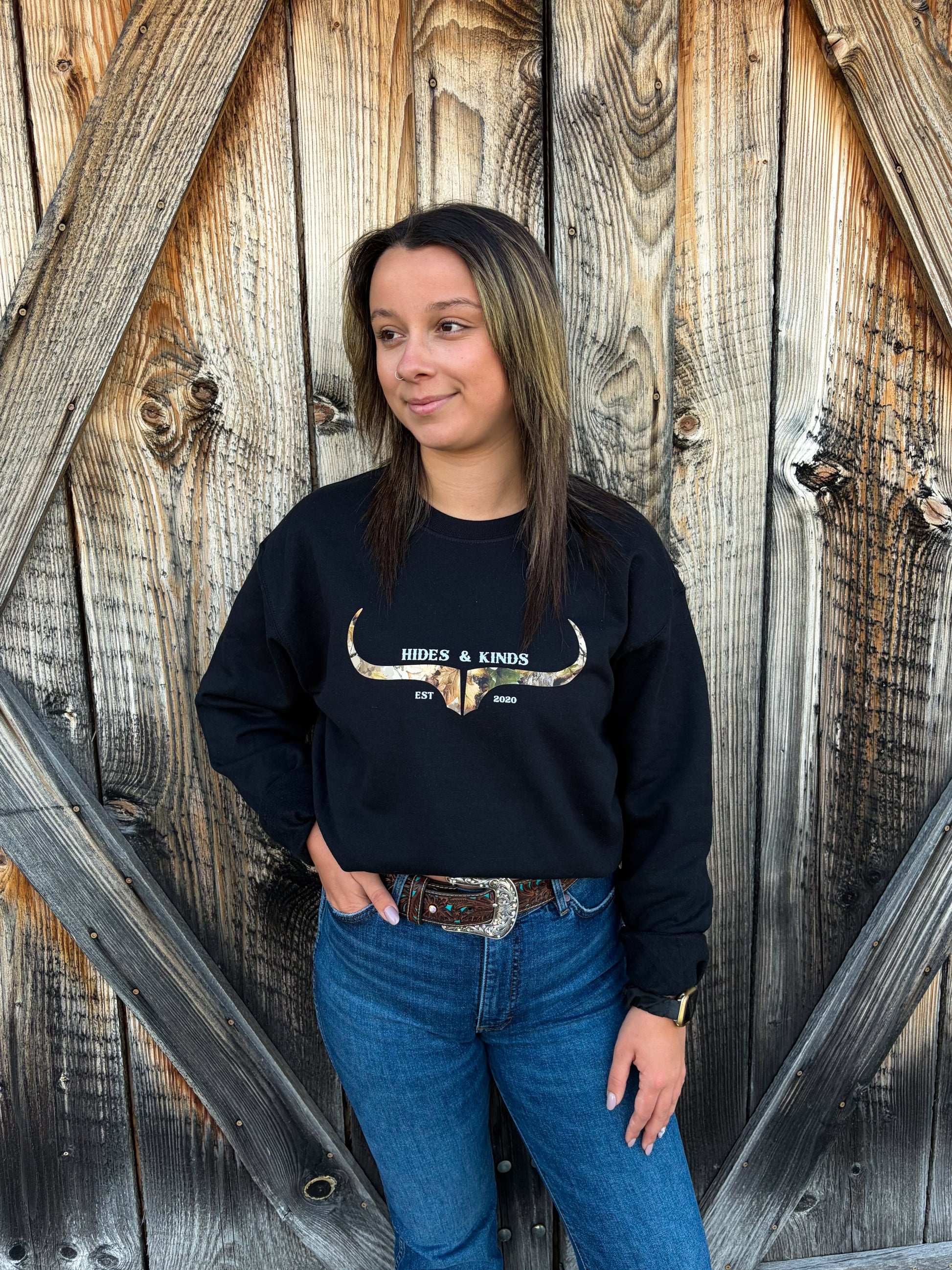 Model wearing the Hides & Kinds Original Logo Crewneck in black with a camouflage longhorn logo, paired with blue jeans in front of a rustic wooden barn door.