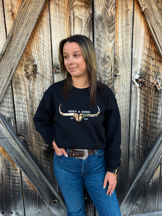 Model wearing the Hides & Kinds Original Logo Crewneck in black with a camouflage longhorn logo, paired with blue jeans in front of a rustic wooden barn door.