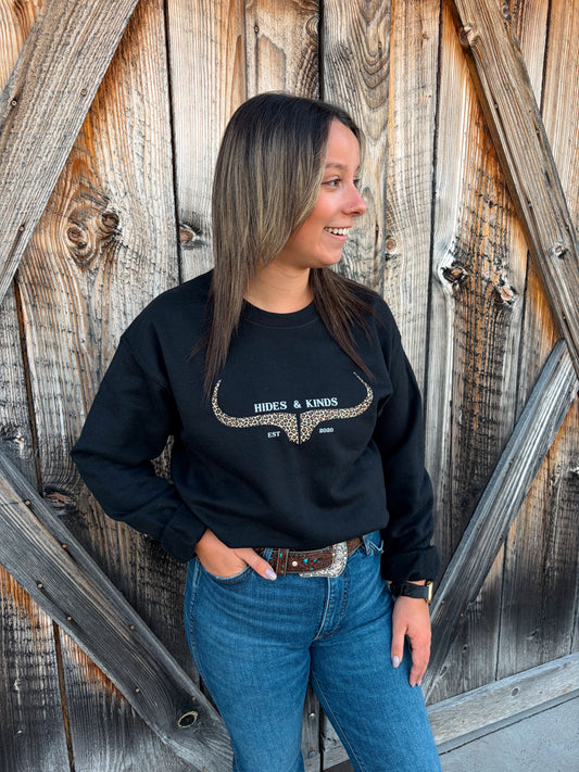 Model wearing the Hides & Kinds Original Logo Leopard Crewneck, a black western sweatshirt with a leopard print longhorn graphic, in front of a rustic wooden barn door.