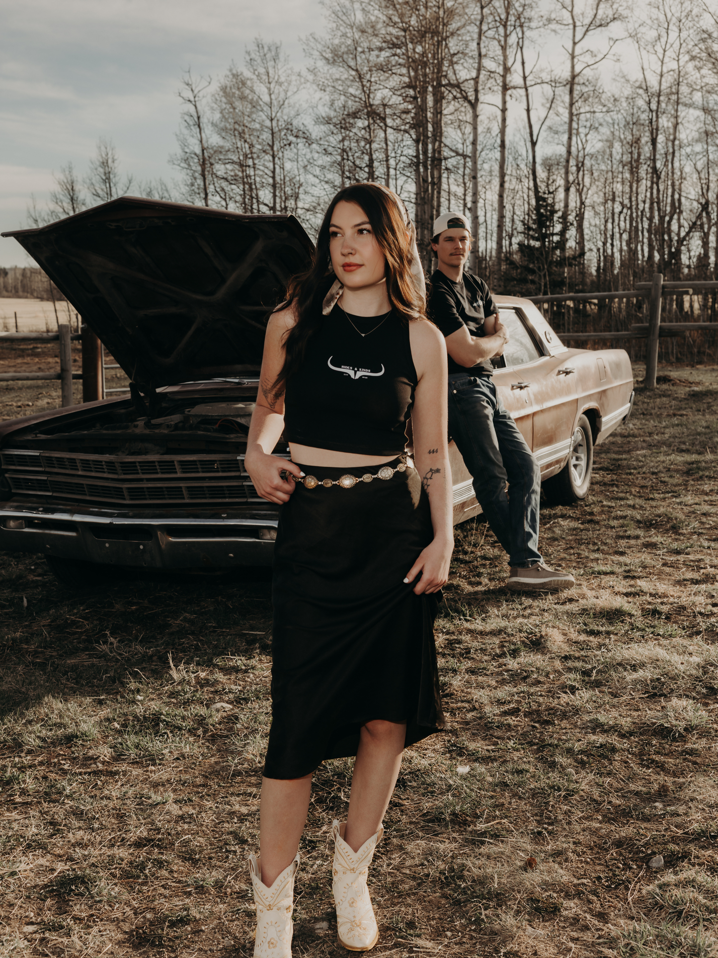Woman wearing a black Hides & Kinds cropped high neck ribbed tank top with white western longhorn logo standing in front of a vintage car in a western inspired photoshoot.