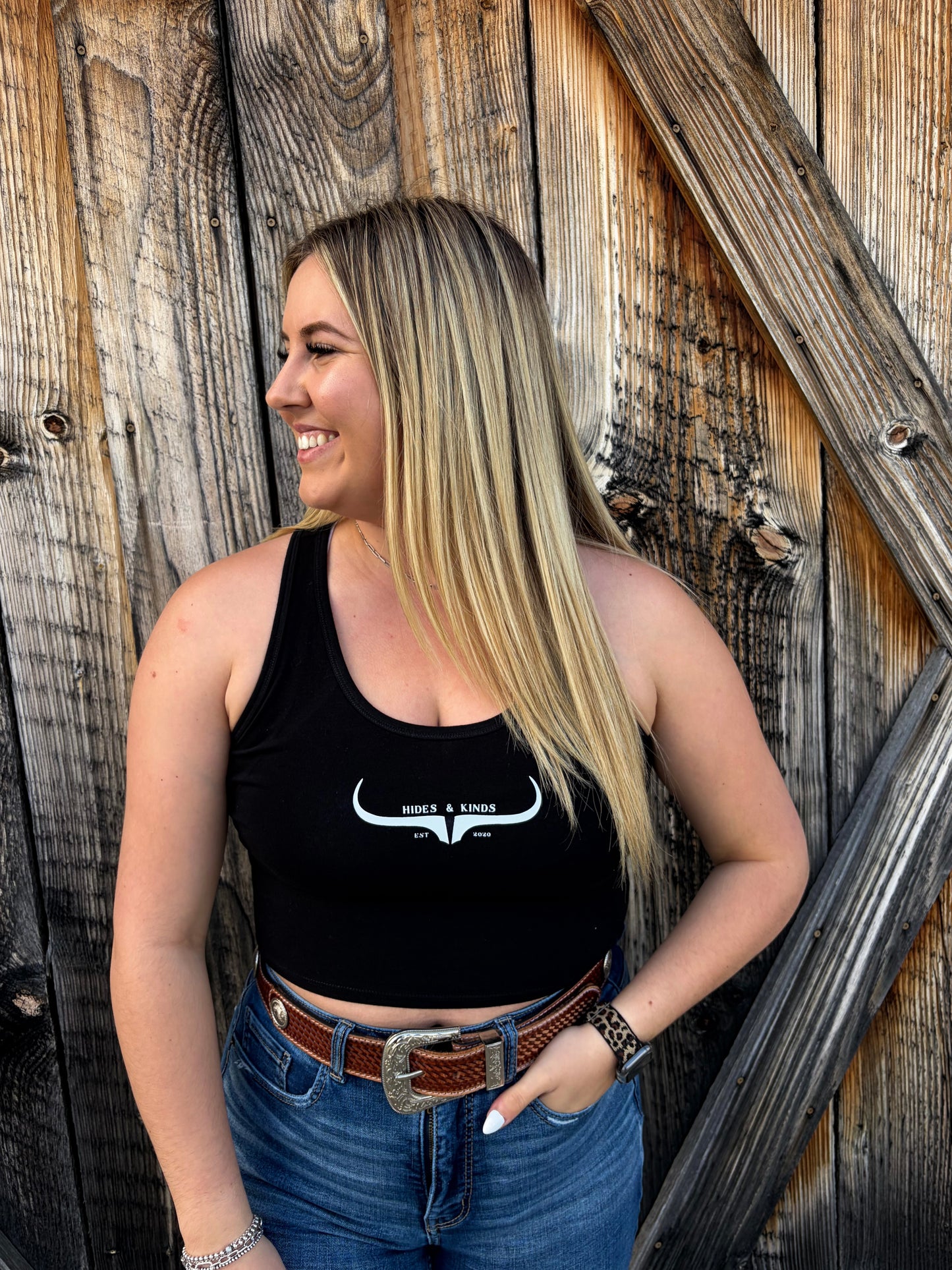 Woman wearing a black Hides & Kinds cropped racerback tank with white western longhorn logo styled with denim jeans and a western belt.