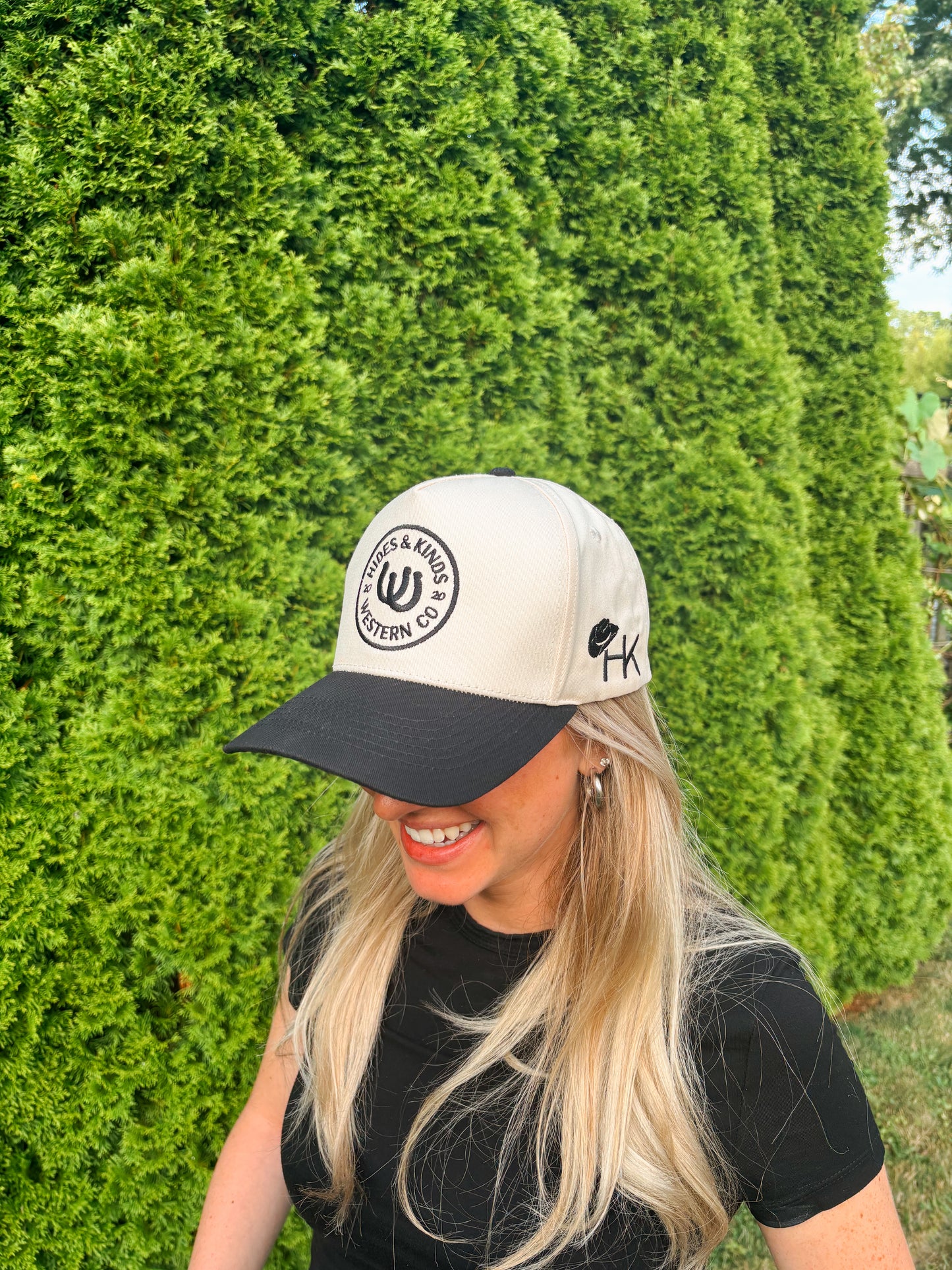 Smiling woman in The Outrider Hat by Hides & Kinds showing logo and side stitching against a green hedge background