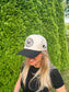 Smiling woman in The Outrider Hat by Hides & Kinds showing logo and side stitching against a green hedge background