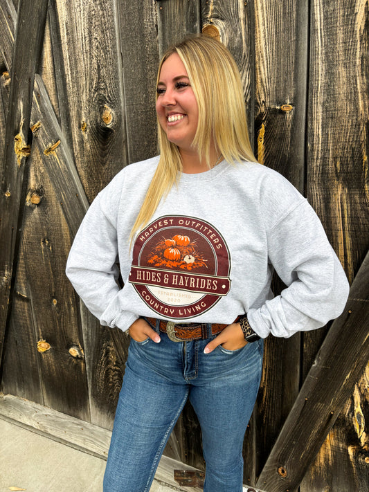 Woman wearing Hides & Hayrides crewneck sweatshirt in front of rustic wood wall, hands in pockets, smiling.