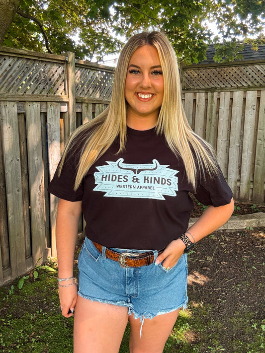 Smiling woman wearing HK Aztec Badge Tee in black with turquoise western logo, styled with denim shorts and a tooled belt in a casual outdoor setting.