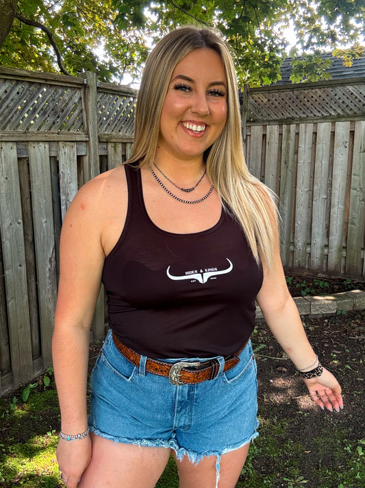 Woman wearing black Hides and Kinds logo tank top with western belt and denim shorts, smiling outdoors.