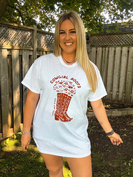 Smiling woman in Hides and Kinds Cowgirl Roots t-shirt with red boot and floral design, styled outdoors.