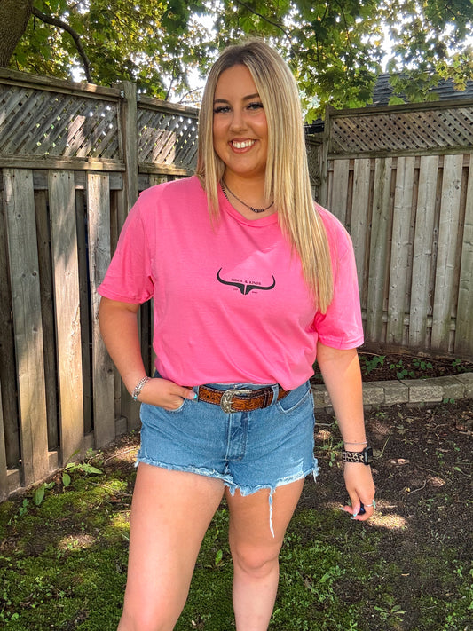 Woman smiling in blossom pink Hides and Kinds logo tee, denim shorts, and tooled leather belt outdoors.