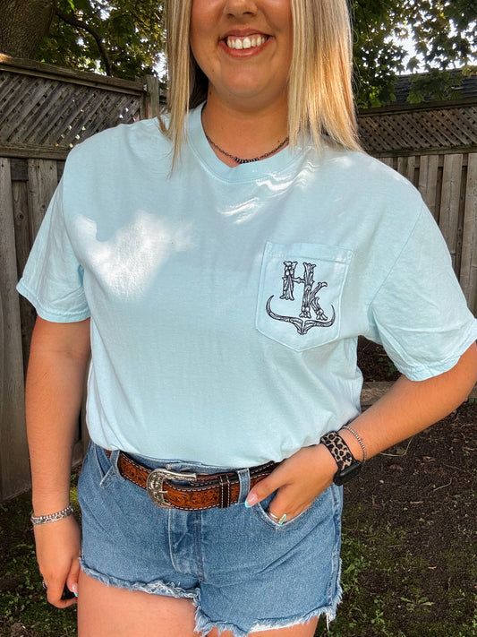 Close-up of Hides and Kinds chambray blue pocket tee with western HK logo design, styled with denim shorts and a tooled belt.
