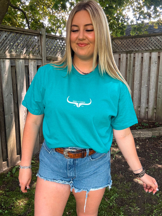 Woman wearing teal Hides and Kinds original logo tee with tooled belt and denim shorts, smiling with a soft closed-eye look.