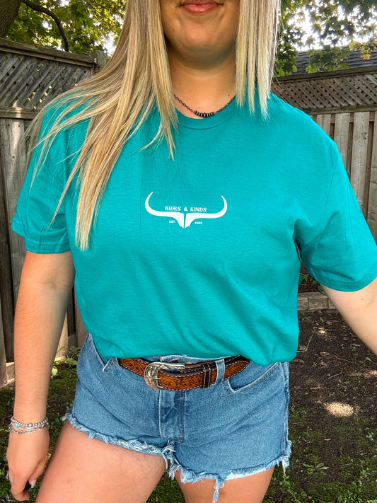 Woman wearing teal Hides and Kinds logo tee styled with western belt and denim shorts, outdoors in natural lighting.