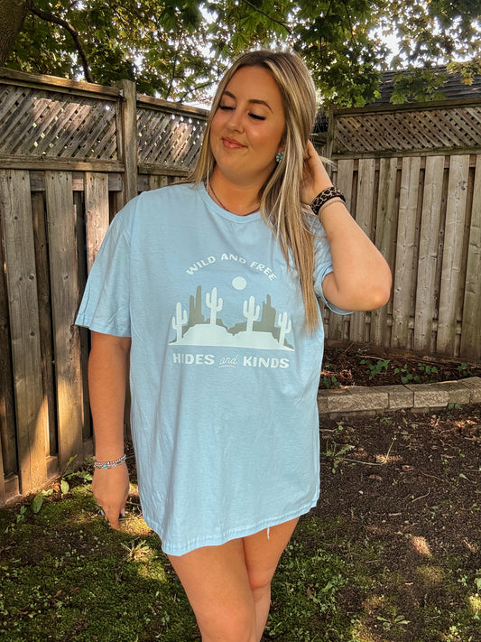 Woman wearing light blue Wild and Free graphic tee by Hides and Kinds with desert landscape print, styled outdoors.