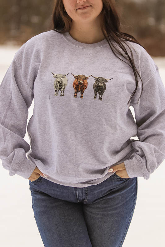 Model wearing heather gray crewneck with three illustrated Highland cows, white, red, and black across the chest.