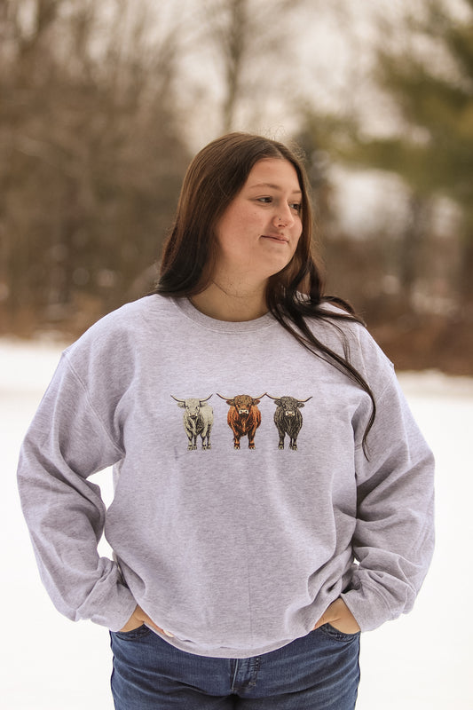 Model smiling in a Highland Herd graphic crewneck, shown in snowy outdoor setting.