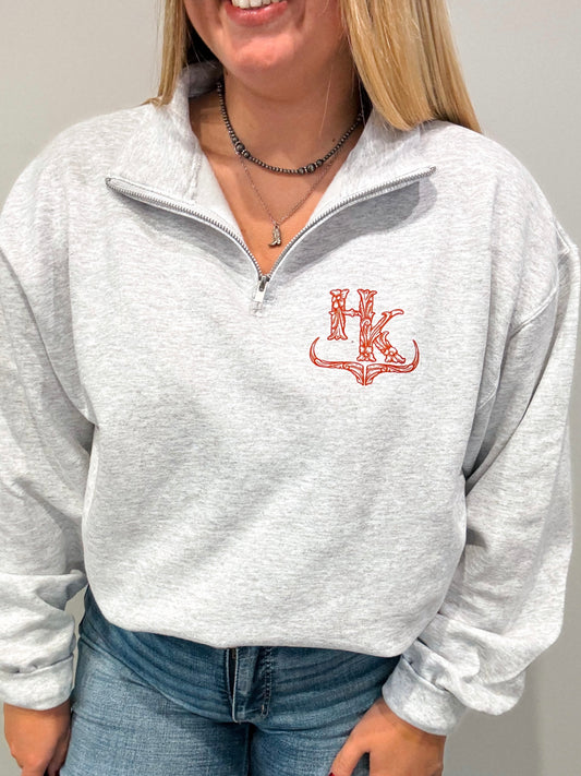Woman wearing the HK Brand Quarter Zip in ash grey with a red logo on the left chest, styled with jeans and layered necklaces.