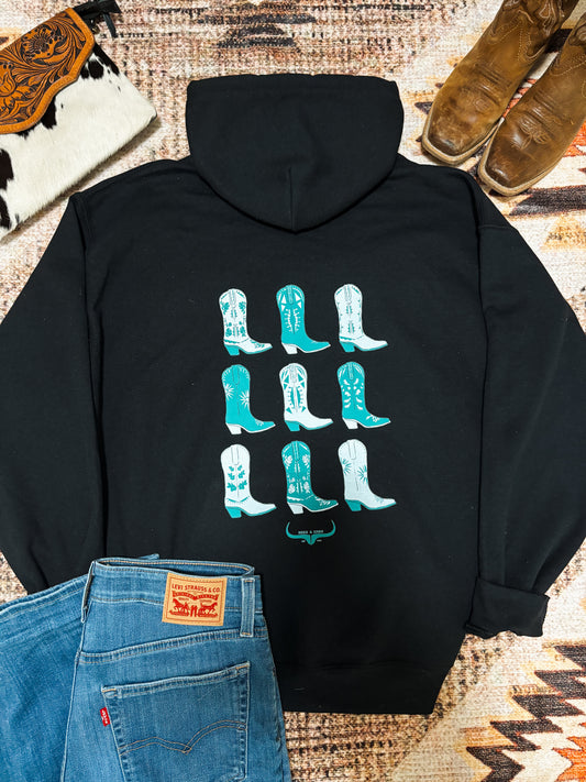 Flat lay of black hoodie featuring nine turquoise western boot graphics on the back, styled with denim, cowhide bag, and brown cowboy boots.