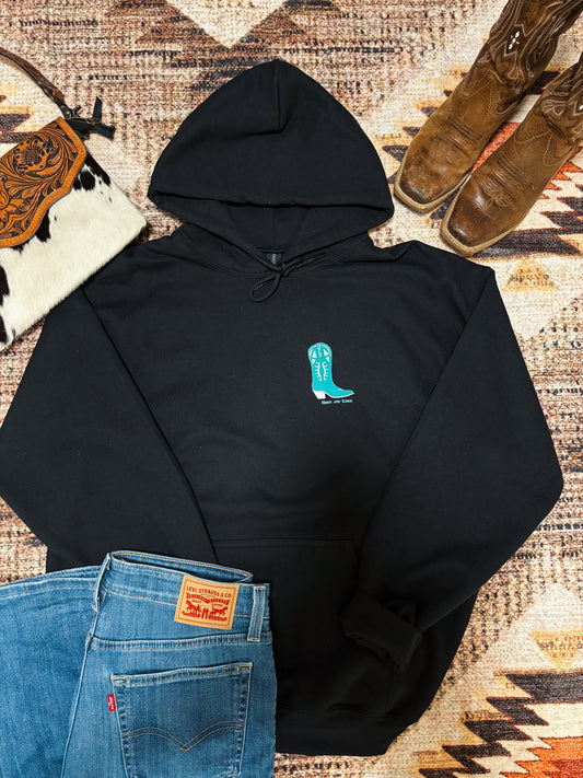 Flat lay of black hoodie with a single turquoise boot graphic on the chest, paired with jeans and rustic western accessories.