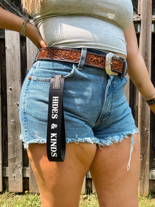 Black branded wristlet with white “Hides & Kinds” text, worn clipped to belt loop with western shorts and belt.