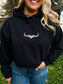 Woman wearing the Hides and Kinds Original Logo Hoodie in black with hands in pockets, showcasing western streetwear style outdoors