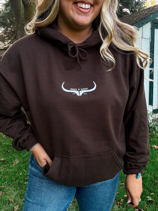 Model wearing the HK Original Logo Hoodie in chocolate brown, smiling with hands in front pocket and standing in a grassy yard.