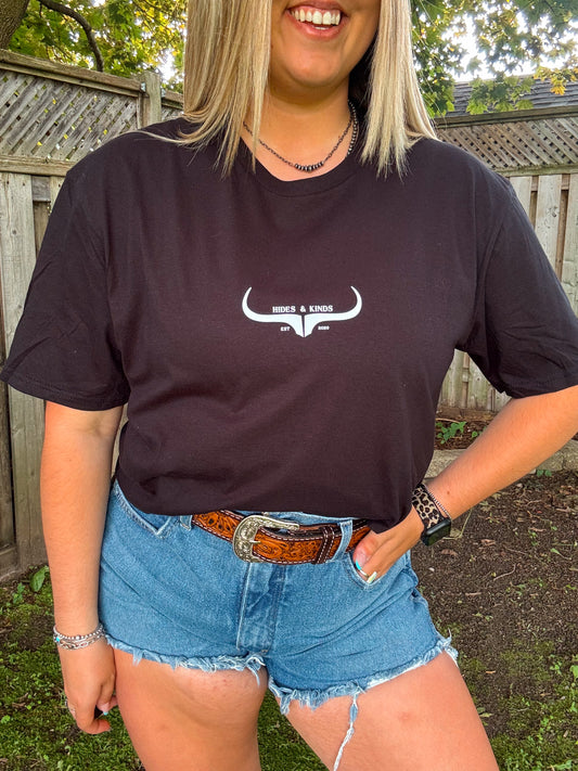 Woman wearing black Hides and Kinds original logo tee paired with a tooled leather belt and denim shorts in a rustic outdoor setting