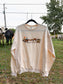 Hides & Kinds Homestead Crewneck in cream hanging on a farm fence with cattle grazing in the pasture behind it.