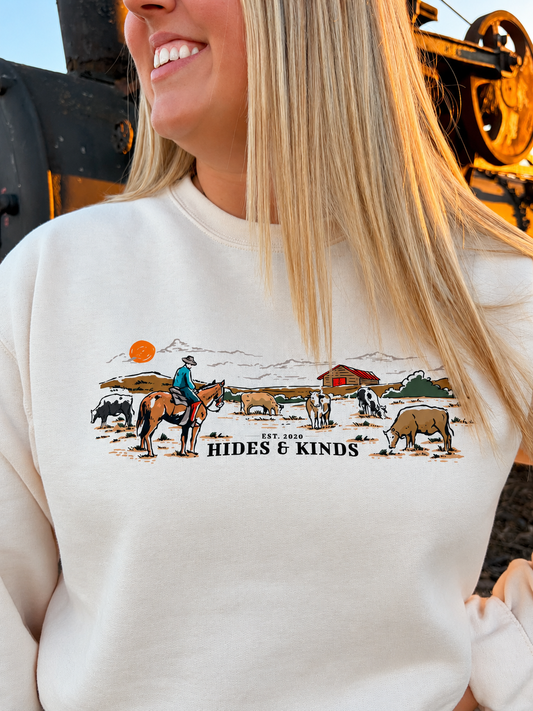 Close-up of the Homestead Crewneck graphic showing a cowboy on horseback, grazing cattle, a red barn and Hides & Kinds branding on cream fabric.