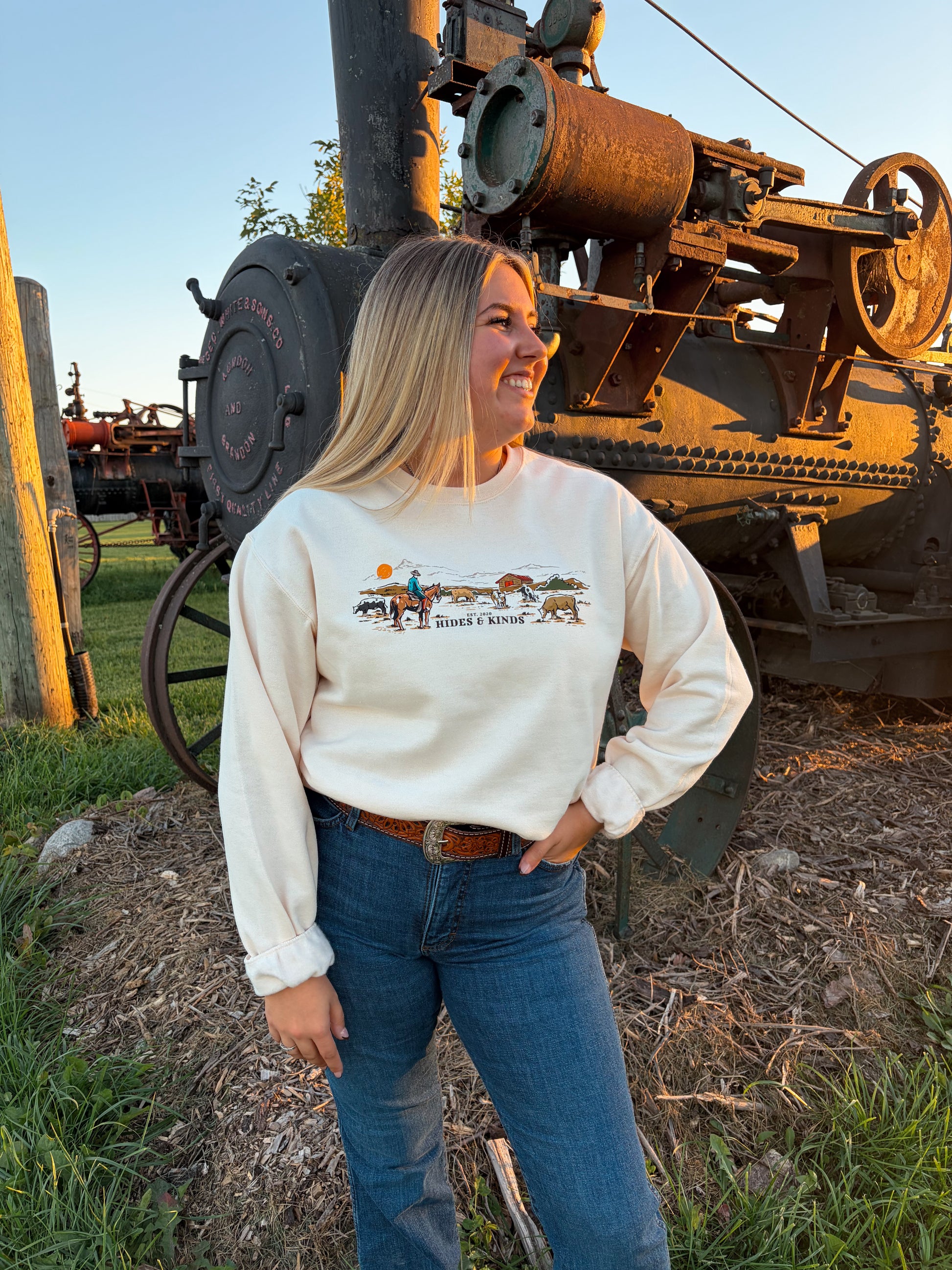 Model styling the cream Hides & Kinds Homestead Crewneck with blue jeans and a western belt outdoors beside antique farm equipment.
