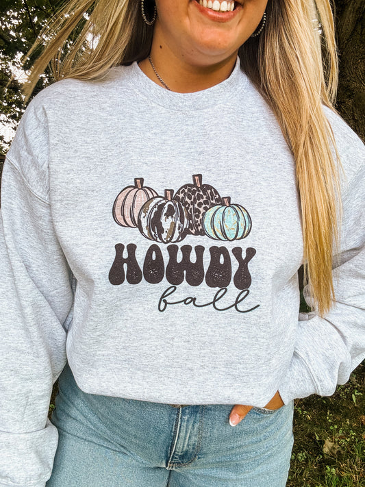 Woman wearing light grey crewneck with cow print and turquoise pumpkins and the text “Howdy Fall,” styled with denim jeans in outdoor setting
