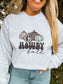 Woman wearing light grey crewneck with cow print and turquoise pumpkins and the text “Howdy Fall,” styled with denim jeans in outdoor setting