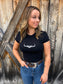 Woman wearing a black Hides & Kinds performance crop top with white western logo, paired with blue jeans and a detailed western belt in front of a rustic wood wall.